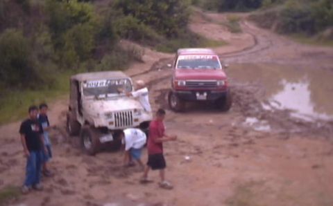 1989 Ford Bronco - Two off-road vehicles navigate a challenging muddy terrain with several people present.
