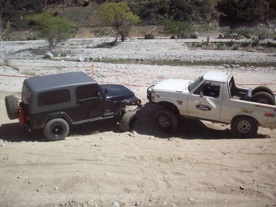 White Ford Bronco with off-road tires beside black Jeep in sandy riverbed