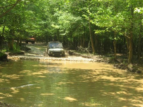 1987 Suzuki Samurai - Two off-road vehicles navigate a muddy water crossing surrounded by dense forest.