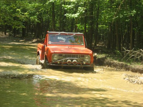 1987 Suzuki Samurai - Orange Ford Bronco navigating a mud pit on a wooded trail.