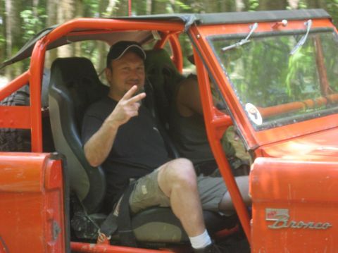 1987 Suzuki Samurai - A man sits in the driver's seat of an orange off-road vehicle, holding up two fingers in a peace sign.