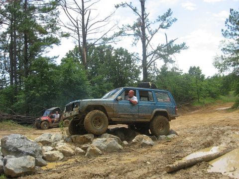 1987 Suzuki Samurai - A blue Jeep Cherokee XJ tackles a challenging rocky obstacle on a muddy off-road trail, with another vehicle visible in the background.
