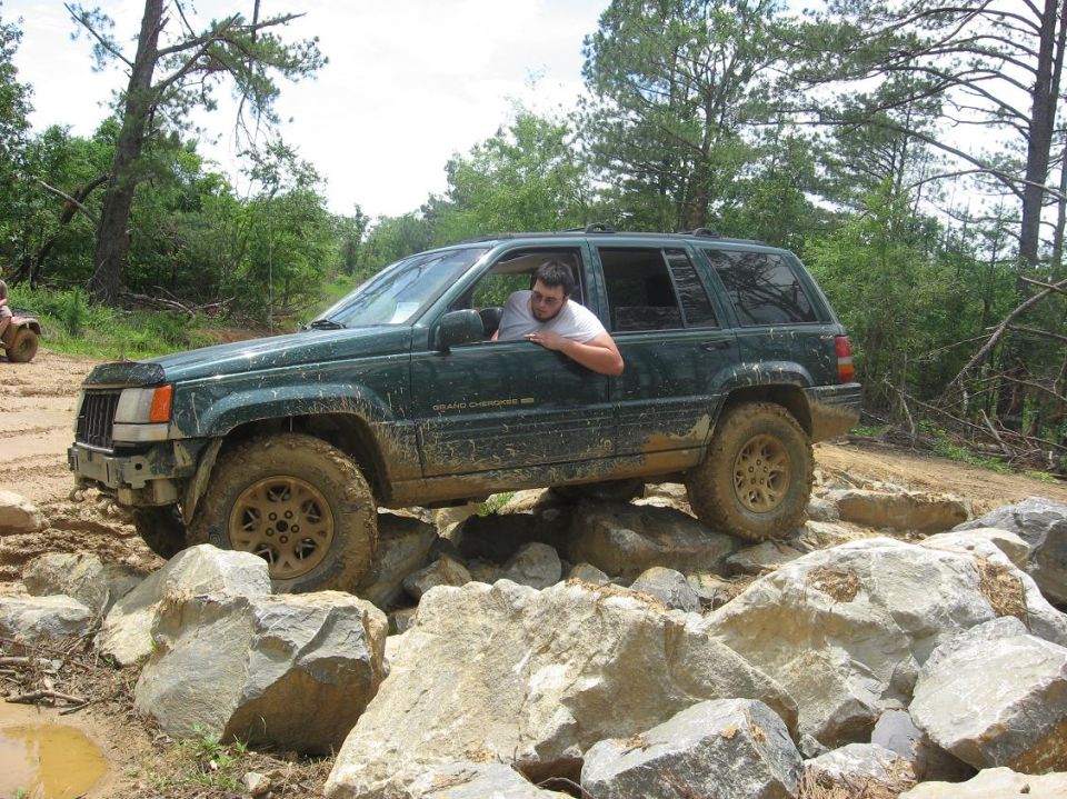 Green Jeep Grand Cherokee navigating large rocks on a trail; another ATV is visible in the background