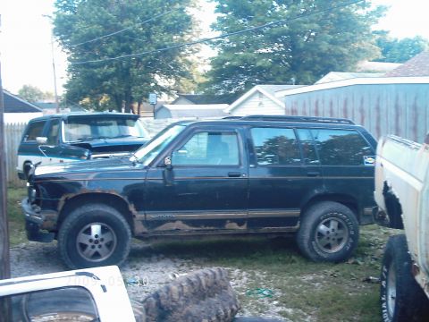 1994 GMC Jimmy - A 1994 GMC Jimmy is seen from the side in a yard, with visible rust on its lower body panels and other vehicles nearby.