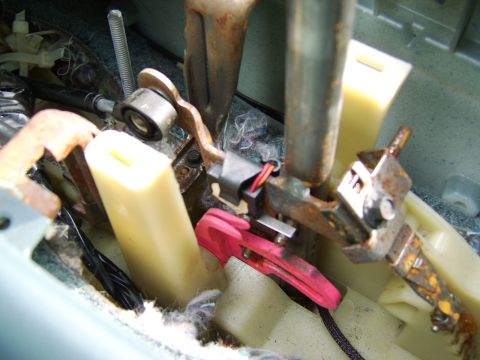 1996 Mercury Grand Marquis - Close-up of a disassembled interior mechanism showing rusty metal brackets, cream-colored plastic components, a threaded rod, a black elect…