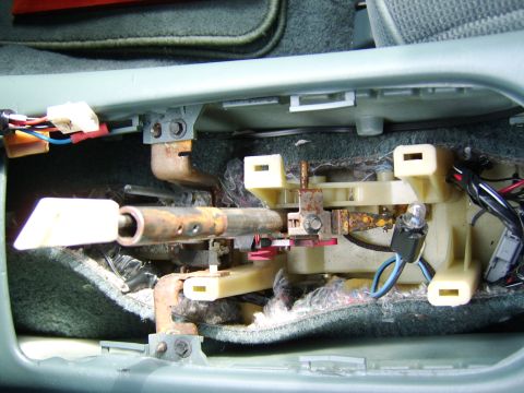 1996 Mercury Grand Marquis - View inside a center console with the trim removed, showing the shifter mechanism, exposed rusty metal linkage, tan plastic housings, and v…