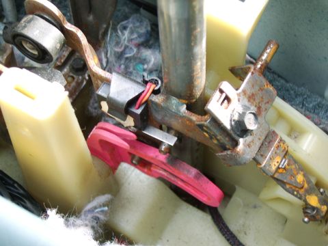 1996 Mercury Grand Marquis - Close-up of a mechanical assembly with rusted steel linkages, cream-colored plastic housings, and a bright pink/magenta lever, along with a…