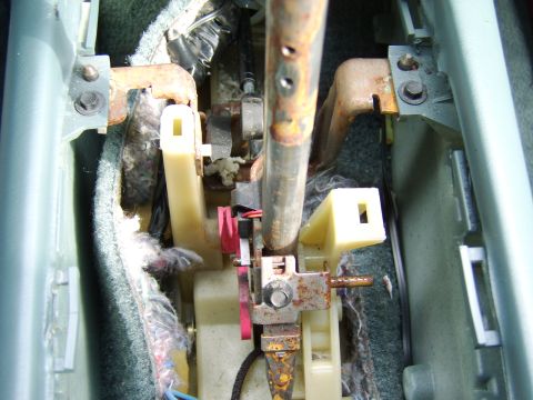 1996 Mercury Grand Marquis - Close-up of an automotive shifter mechanism viewed from above through an opening in gray carpet and trim, showing a rusty steel shaft, moun…
