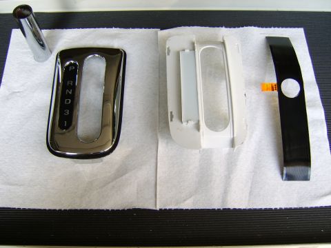 1996 Mercury Grand Marquis - Three automatic shifter bezel/trim components laid out on white paper over a black ribbed mat: a chrome PRND321 gate trim on the left, a wh…