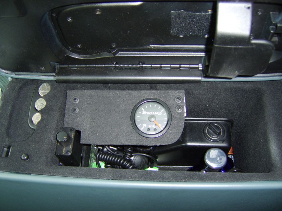 Vacuum gauge mounted inside a car's center console storage compartment with wiring and coins.