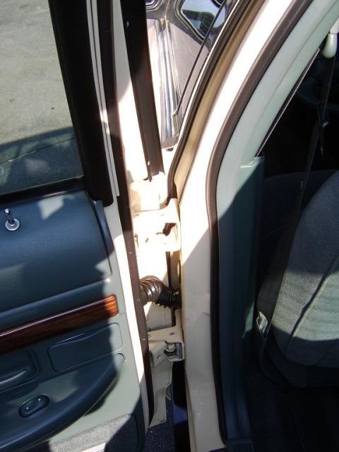 1996 Mercury Grand Marquis - Close-up of an open front door jamb showing the exposed door hinges, wiring conduit and body-side pillar of a tan/beige sedan, with the gra…