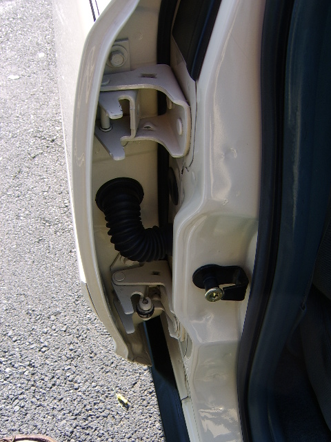 1996 Mercury Grand Marquis - Close-up of the front door jamb area on a beige/cream vehicle, showing the exposed upper door hinge bracket, the black accordion-style wiri…
