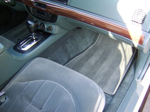 1996 Mercury Grand Marquis - Interior view of the passenger-side front floor area, showing gray carpeted floor mats, the center console with a chrome-trimmed automatic…