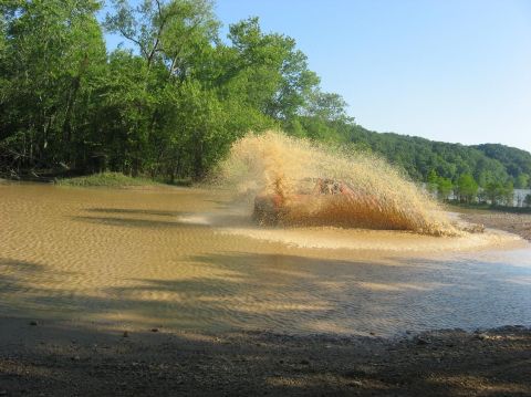 1987 Suzuki Samurai - A vehicle powers through a muddy water obstacle, creating a large splash.