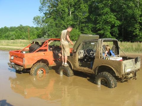 1987 Suzuki Samurai - Two modified off-road vehicles are stuck deep in a muddy puddle on a sunny day.