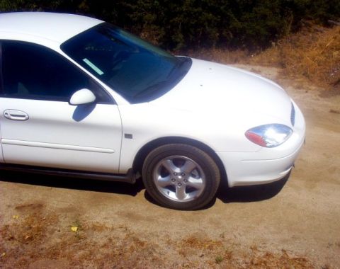 2000 Ford Taurus - Front passenger side of a white sedan parked outdoors on a dirt surface with vegetation in the background.
