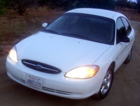 2000 Ford Taurus - A white 2000 Ford Taurus is parked on a dirt road with its headlights on.