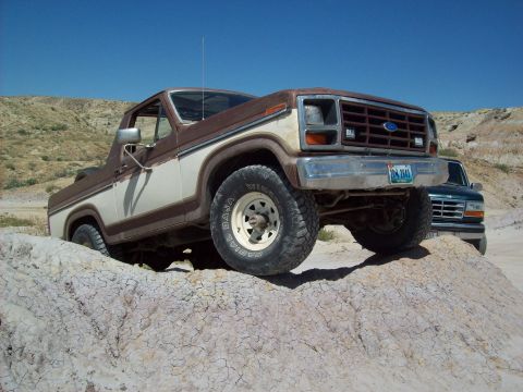 1985 Ford Bronco - A two-tone Ford Bronco navigates off-road terrain, with another vehicle visible in the background.