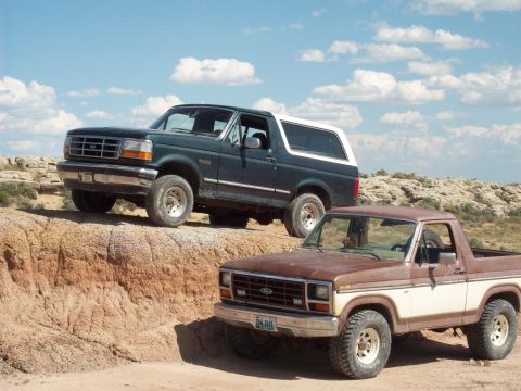 1985 Ford Bronco - Two Ford Broncos, a brown and white model in the foreground and a dark green one behind it, navigate a dirt trail under a cloudy sky.