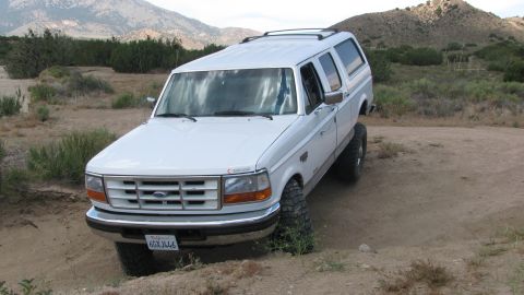 1996 Ford C-350 (Centurion Conversions) - A white Ford SUV with a camper shell is parked on a dirt road with hills in the background.