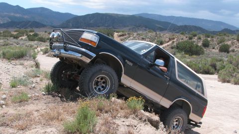1996 Ford C-350 (Centurion Conversions) - A dark green and tan two-tone Ford Bronco flexing its front suspension as it climbs a rocky dirt embankment, front driver-side wheel lifted…