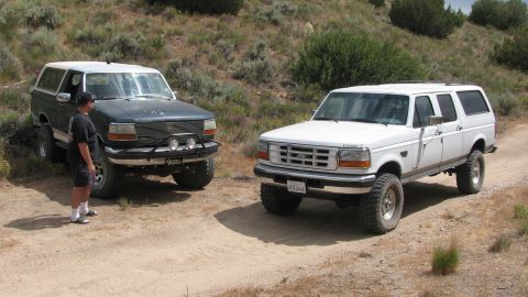 1996 Ford C-350 (Centurion Conversions) - Two modified Ford SUVs, a white Centurion conversion and a dark green Bronco, are parked on a dusty, off-road trail.