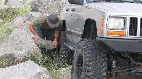 1996 Ford C-350 (Centurion Conversions) - A man adjusts a rock near the front tire of a silver Jeep Cherokee XJ during an off-roading excursion.