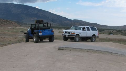 1996 Ford C-350 (Centurion Conversions) - Two off-road vehicles are parked on a dirt path with a mountainous landscape behind them.