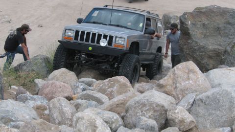 1996 Ford C-350 (Centurion Conversions) - A silver Jeep Cherokee XJ is driven over large rocks, with people nearby.