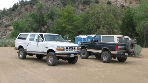 1996 Ford C-350 (Centurion Conversions) - Two lifted Ford SUVs parked nose-to-tail on a sandy staging area: a white two-door Bronco-style rig facing left on chrome-modular wheels an…