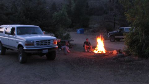 1996 Ford C-350 (Centurion Conversions) - Dusk campsite scene with a white 1990s Ford crew-cab conversion SUV parked at left in 3/4-front view on lifted suspension and chunky tires,…