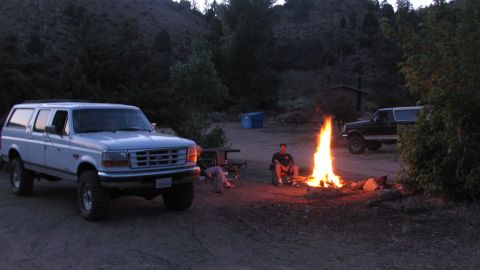 1996 Ford C-350 (Centurion Conversions) - A white lifted 1990s Ford crew-cab pickup with a matching bed-height camper shell sits parked at a dirt campsite at dusk, wearing oversized…