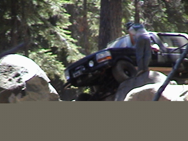 Dark blue Ford Bronco off-roading up large boulders in a forest with a spotter standing nearby
