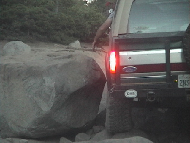 Rear of a tan Ford SUV with brake light on, squeezing past a large boulder on a rocky off-road trail