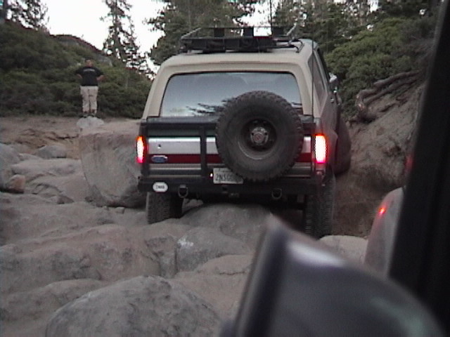 Rear view of tan Ford Bronco with spare tire and roof rack crawling over boulders on a rocky trail