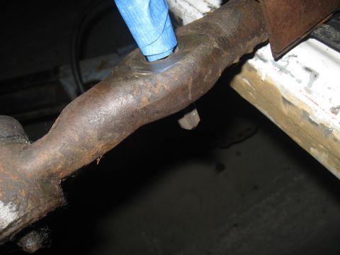 1978 Ford Bronco - Close-up view of a heavily rusted metal linkage, with blue tape obscuring a hole. The part appears to be from a vehicle's undercarriage.