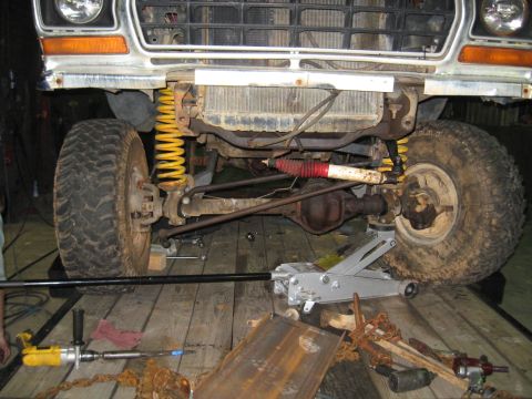 1978 Ford Bronco - The front suspension and drivetrain of a vehicle are visible, with a jack supporting the frame and tools scattered around.