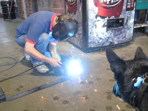 1978 Ford Bronco - A man welds a metal bar while a dog watches, with a vending machine in the background.