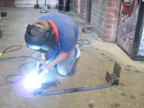 1978 Ford Bronco - A person in a welding mask and jeans is kneeling and welding a metal frame on a concrete floor.
