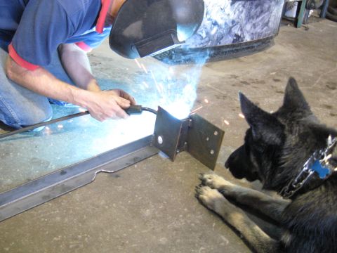 1978 Ford Bronco - A man welds a metal bracket on a concrete floor, with a German Shepherd lying nearby.