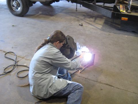 1978 Ford Bronco - Welding a metal part indoors with a trailer and truck nearby.