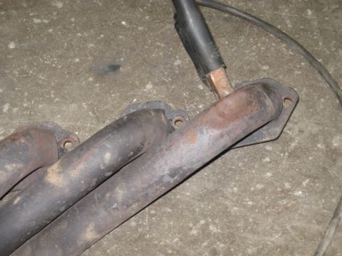 1978 Ford Bronco - This image shows a close-up of a rusty exhaust manifold with a welding cable connected to it.
