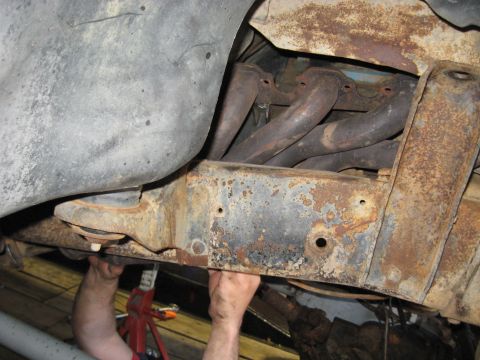 1978 Ford Bronco - Mechanic's hands are visible working on the underside of a vehicle, focusing on the rusty frame and exhaust manifold.
