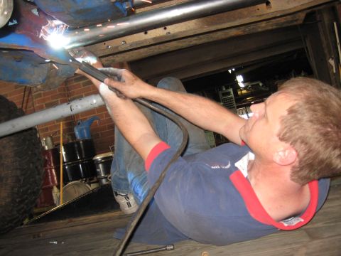 1978 Ford Bronco - A person is welding a pipe underneath a vehicle, with a large tire and storage drums in the background.