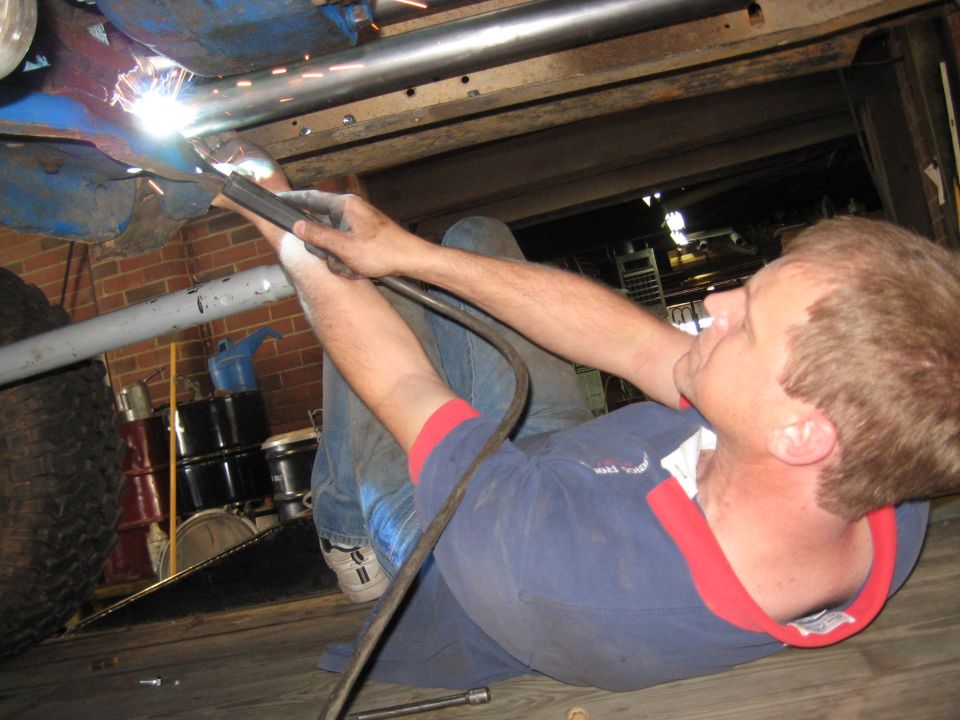 guy welding exhaust pipe under a vehicle, large tire visible