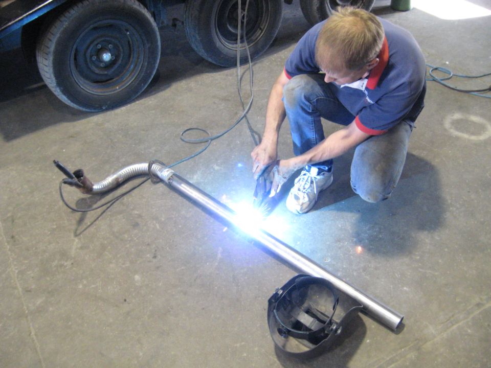 Man welding a metal exhaust pipe with a flexible section, wearing jeans and a blue shirt. Welding mask on the floor