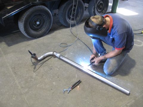 1978 Ford Bronco - A person is welding a section of exhaust pipe on a concrete floor, with a trailer visible in the background.