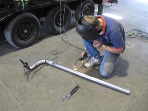 1978 Ford Bronco - A person welds an exhaust pipe on a concrete floor, with a trailer in the background.