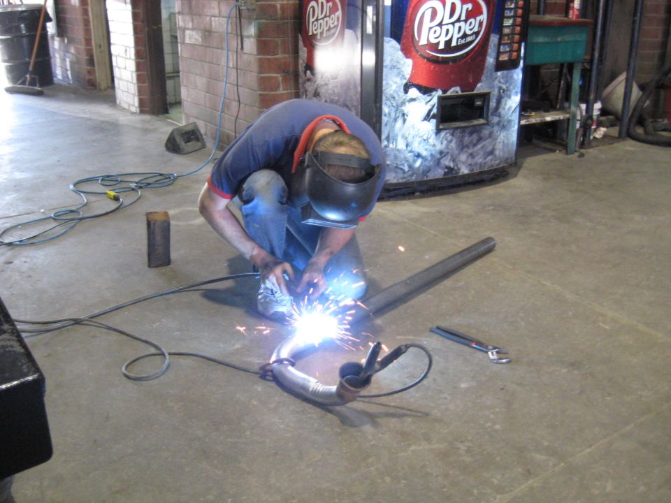 Welder sparks fly as exhaust pipe is welded. Dr Pepper vending machine in background