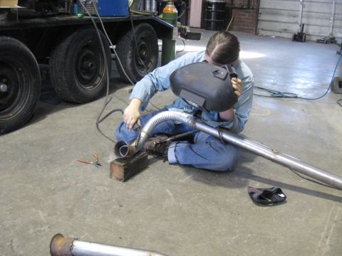 1978 Ford Bronco - A person welds an exhaust pipe in a workshop setting, with a trailer and tools in the background.
